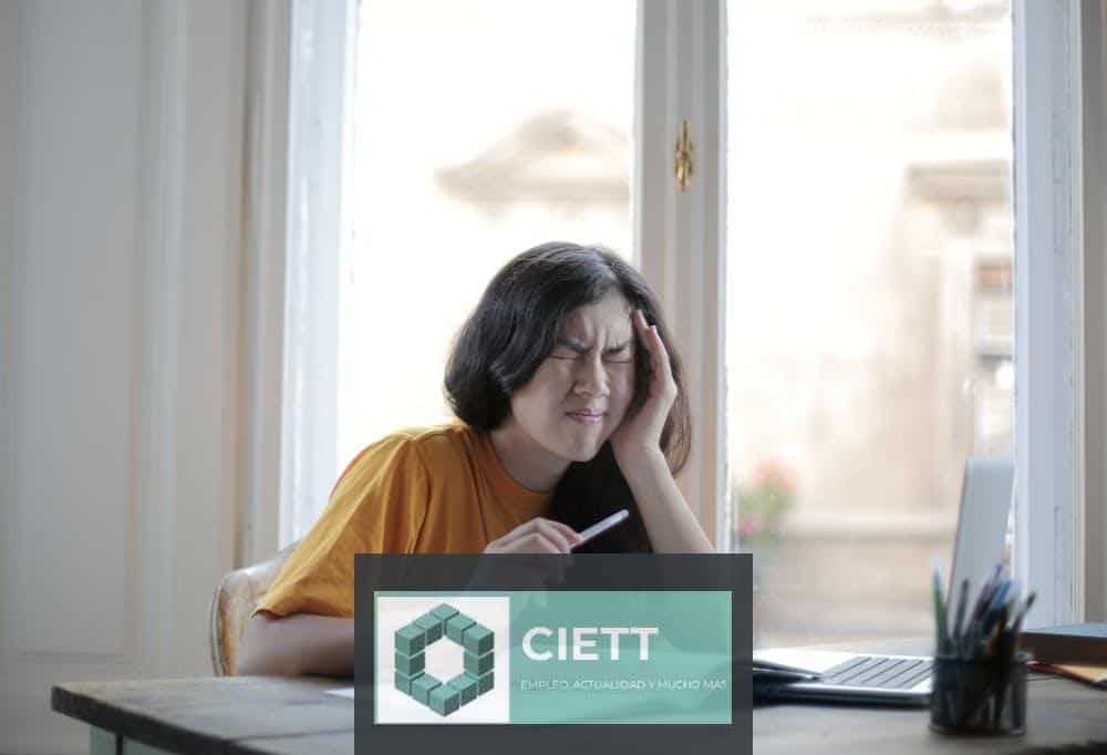 Ethnic female student in casual wear sitting at table with laptop and notebook and grimacing from migraine while doing homework during distance learning