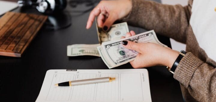 Woman counting cash at desk with notebook, pen, and headphones nearby.